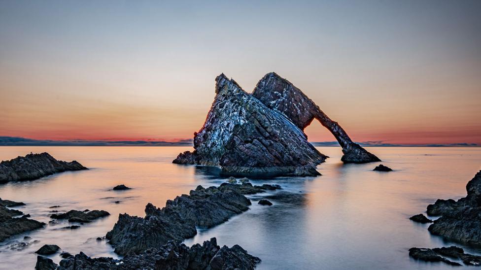A large natural sea arch rises from calm water at sunrise or sunset. Jagged rocks surround the formation, while soft orange and pink colours glow along the horizon beneath a clear sky.