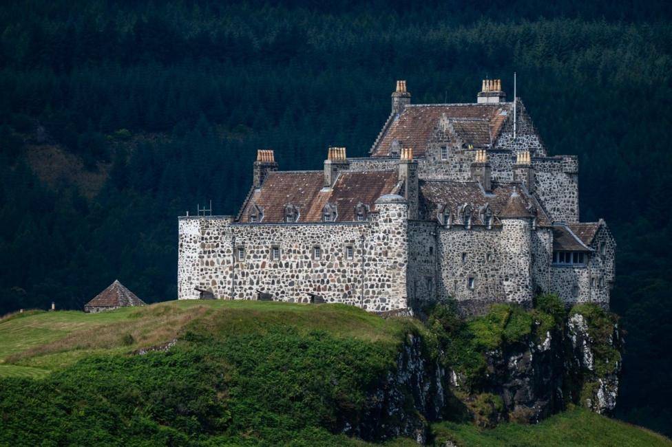A large stone castle stands on a rocky cliff surrounded by green countryside. The historic building features thick stone walls, multiple chimneys and steep roofs, with a dense forest forming a dark backdrop.