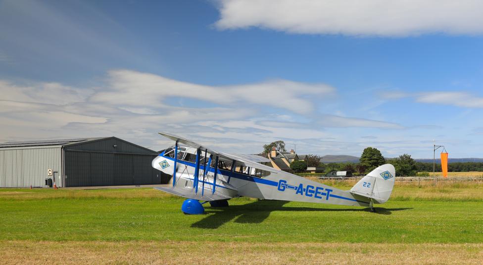 A blue-and-white vintage biplane sits on a grassy airfield beside a large hangar. The aircraft is parked under a bright blue sky with scattered clouds, with countryside and buildings visible in the background.