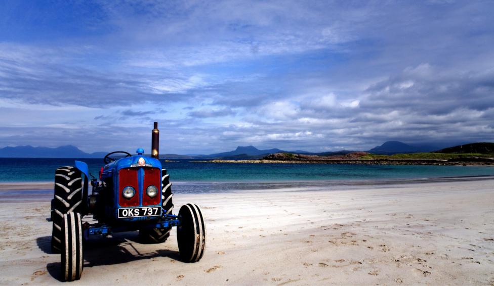 A blue vintage tractor stands on a wide white-sand beach beside turquoise water. Low rocky islands and dark mountains are visible in the distance beneath a dramatic sky filled with thick grey and blue clouds.