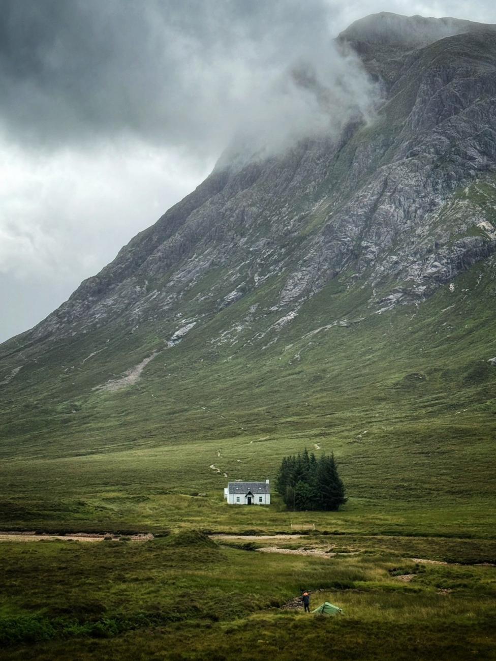 A small white cottage sits alone on a wide green moor beneath a towering rocky mountain. Low cloud and mist cling to the upper slopes, while a winding path leads across the landscape toward the house. In the foreground, a small tent and a person stand near a stream.