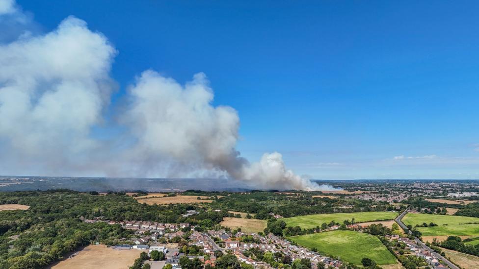 A bird's eye drone view of fields, trees and houses. In the background white smoke billows into the sky. The sky is blue.