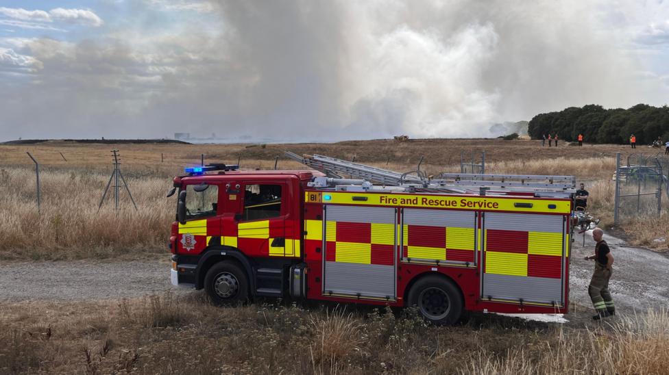 A red and yellow fire engine is parked up on a dirt track, with dry brown grass either side. In the background is white smoke billowing.
