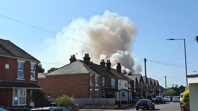 White smoke billowing above a row of bricked terraced houses, with cars parked on the road. The sky above is blue.