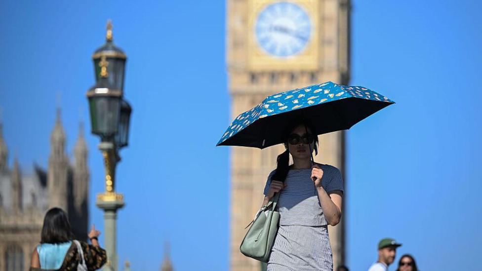 A woman walks in the shade of an umbrella in front of Big Ben in London, with clear blue skies overhead