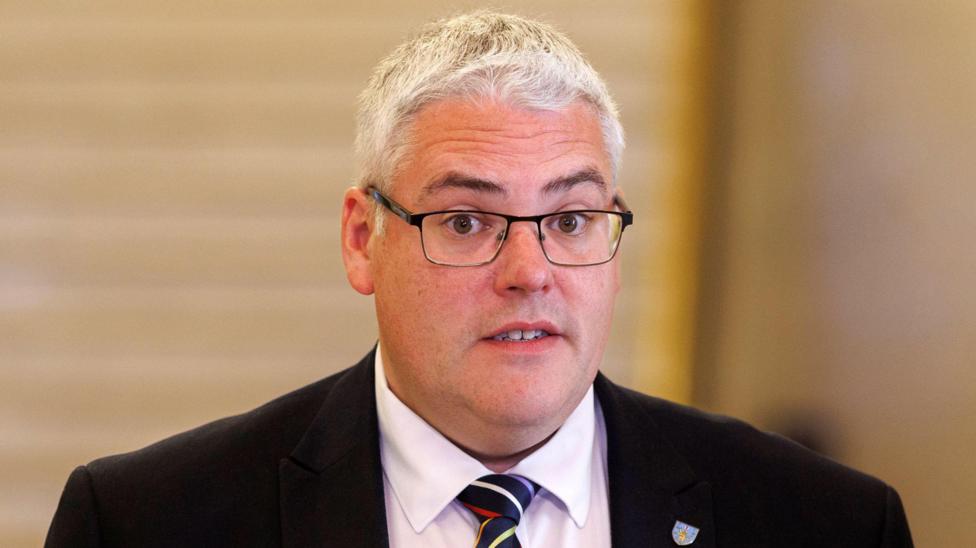 Gavin Robinson, dark suit, stripy tie. He has glasses and grey hair