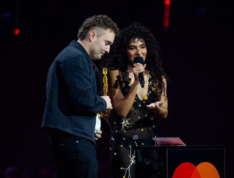 Picture of Sam Fender in a blue jacket, holding a Brit award as Olivia Dean speaks into a microphone on stage wearing a black and yellow dress