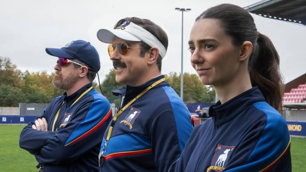 Three people are seen dressed in blue sports gear. The man on the left has a blue baseball gap and sunglasses, the man in the middle has a white visor, sunglasses and moustache, and on the end is a woman with brown hair tied back. They are standing in a football stadium