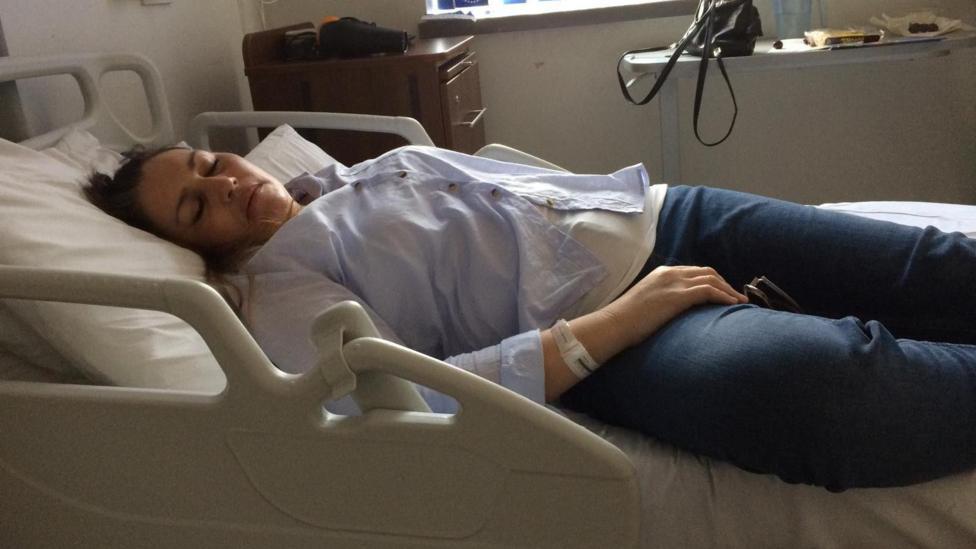 Victoria lying down flat on a hospital bed sleeping, next to a chest of drawers.