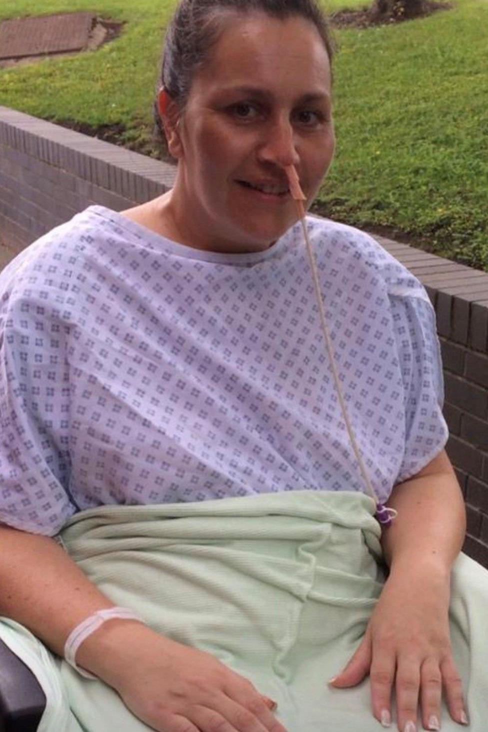 Victoria wearing a purple hospital gown while sitting in a wheelchair, and has a piece of equipment inserted in one of her nostrils. She is looking at the camera.