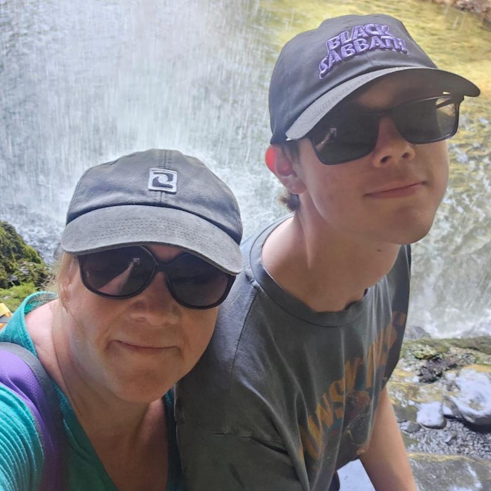 A mother and son wearing baseball caps and sunglasses and standing in front of a waterfall