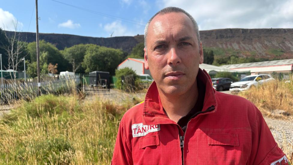 Craig, a man with very short hair is wearing a red jacket with the collar turned up. In the background there's an industrial-looking building and beyond that is a mountainside which has been partially burnt.