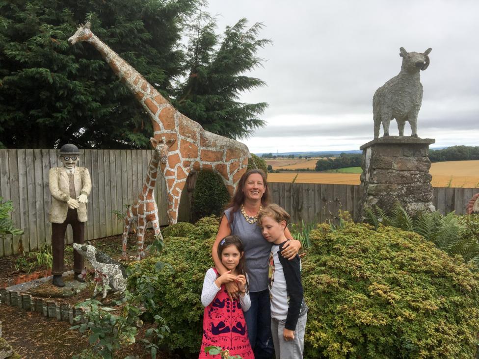 A woman with her arms around two children, standing in a rural garden. Behind her are statues of animals including two giraffes.