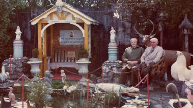 Two men with grey hair sit among a garden with dozens of cement animals and creatures
