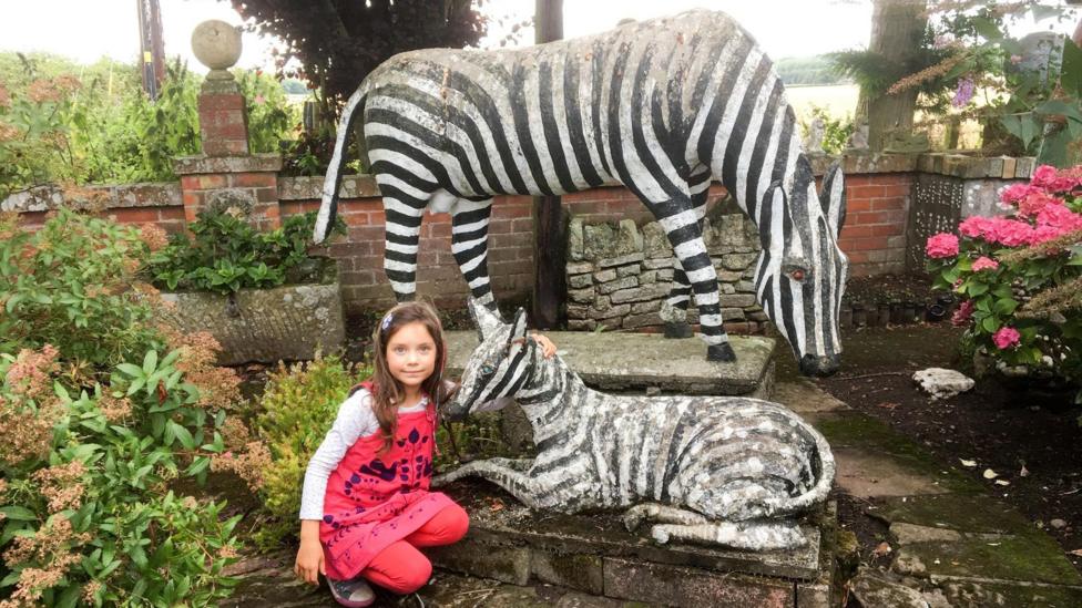 A little girl with long brown hair in a pink top and trousers crouches down next to a statue of a zebra with another larger zebra statue behind her