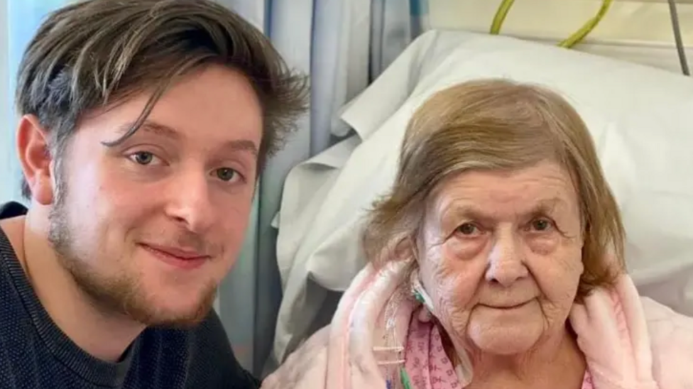 A young man in a blue t-shirt is smiling and crouching next to an older woman in a hospital bed wearing a pink dressing gown.
