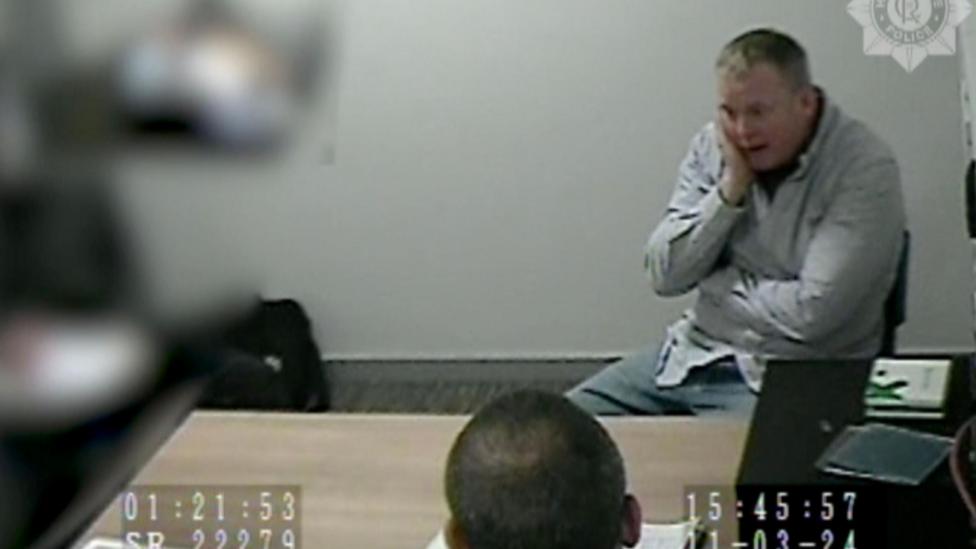 A man in a grey shirt is sitting behind a desk with his hand pressed to his face. The back of the head of a second man is in the foreground. The image is stamped with the logo of Humberside Police