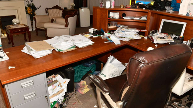 A brown leather chair is in front of a wooden desk piled with paperwork. Under the desk the bin is overflowing with paper. There are two leather chairs, table and a fireplace in the background.