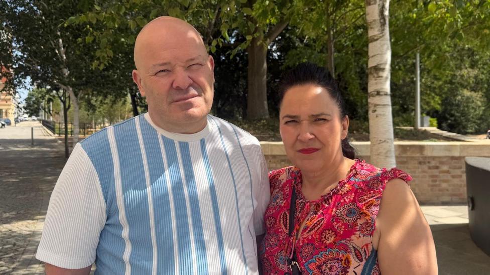 A man and a woman stand together on a pavement in front of a public garden. They have serious expressions on their faces. The man has a shaved head and wears a T-shirt with blue and white vertical stripes. The woman on the right has black hair, tied back and wears a red, pattered top.