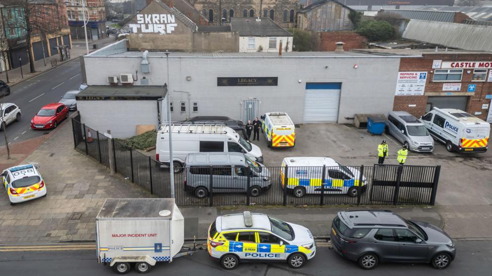 A number of police cars and vans are surrounding a large grey warehouse which has the sign saying Legacy on its wall