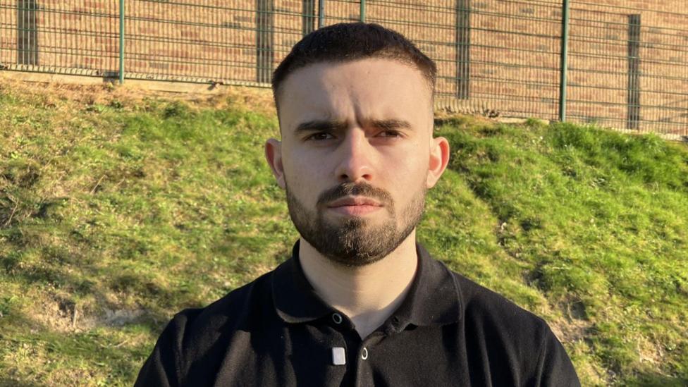 George is standing outside by some green playing fields and looking directly at the camera. He has short dark hair and a neatly clipped dark beard. He has dark eyes and is wearing a black polo shirt. He looks serious.