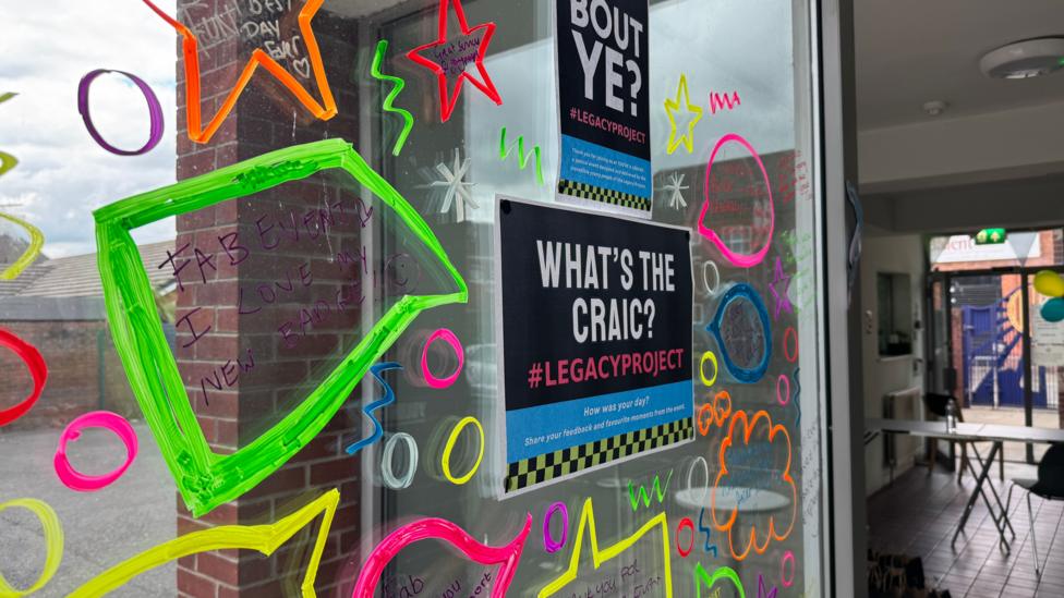 A glass window is covered in bright doodles, shapes and different written messages in coloured pen. In the middle there are two printed signs from the Legacy Project with one titled what's the craic and the other saying Bout Ye.