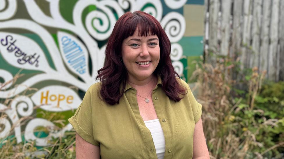 Siobhan McKinley is a young woman with shoulder-length hair which has been dyed a dark read colour. She is smiling at the camera. She is wearing a green dress with a white shirt underneath and a silver necklace. She is standing in front of some bushes and a large mural which has strength, hope and community written on the trees.