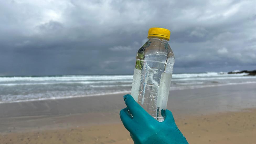 A hand - wearing a protective glove that is green - is holding up a plastic bottle containing water at a beach. It is a sample of the bathing water, there is golden sand and breaking waves in the background. The sky is cloudy.
