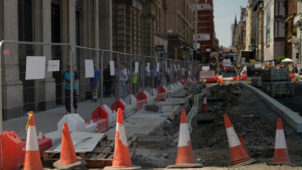 Roadworks along a city centre street with traffic cones, safety barriers and fenced-off excavations. Pedestrians walk along a temporary pavement beside construction works, with buildings and traffic visible in the background.