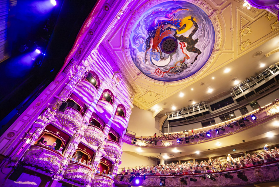 The restored auditorium of Edinburgh's King's Theatre is illuminated in purple and blue light as audience members fill the stalls, balconies and upper circle. The theatre's ornate decorative interior and the dome painting All the World's a Stage by John Byrne are visible above the auditorium.