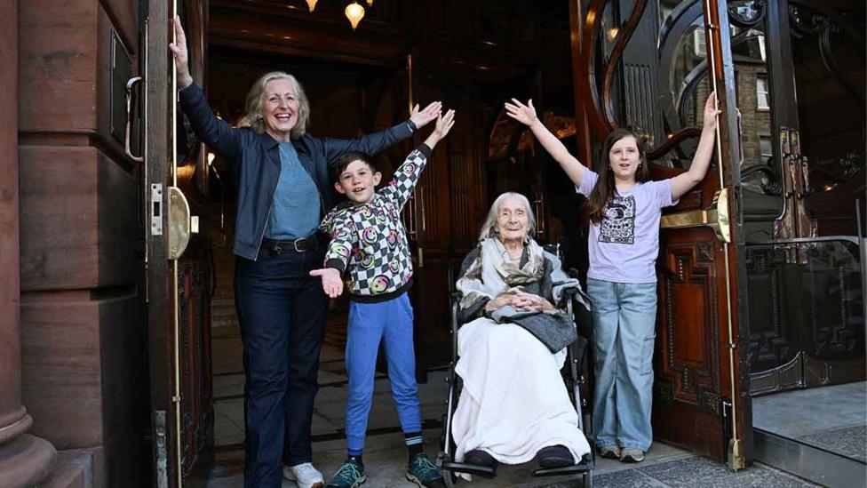 Four people in the open doorway of a theatre - a woman, a boy and a girl standing with their arms held aloft, and an older woman sitting in a wheelchair smiling.