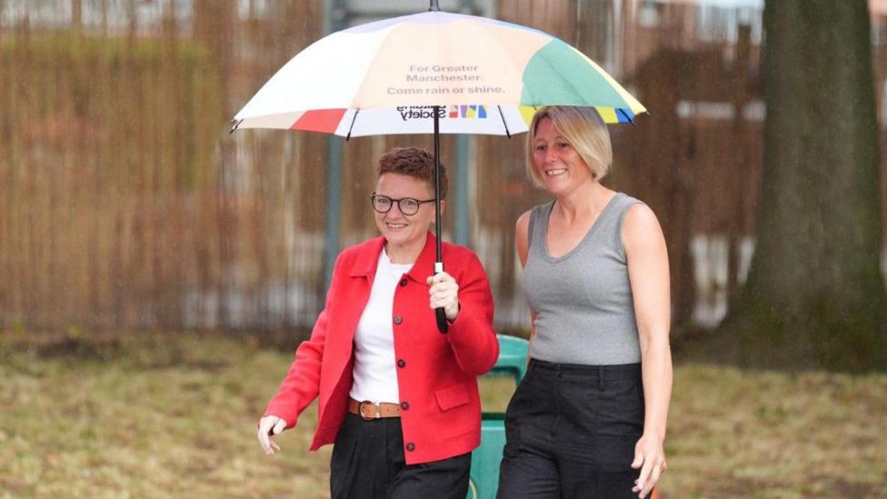 Labour's Bev Craig arrives at Broad Oak Primary in Didsbury with her partner Tracy to vote in the Greater Manchester mayoral by-election. She holding a multicolour umbrella as she walks towards the polling station.