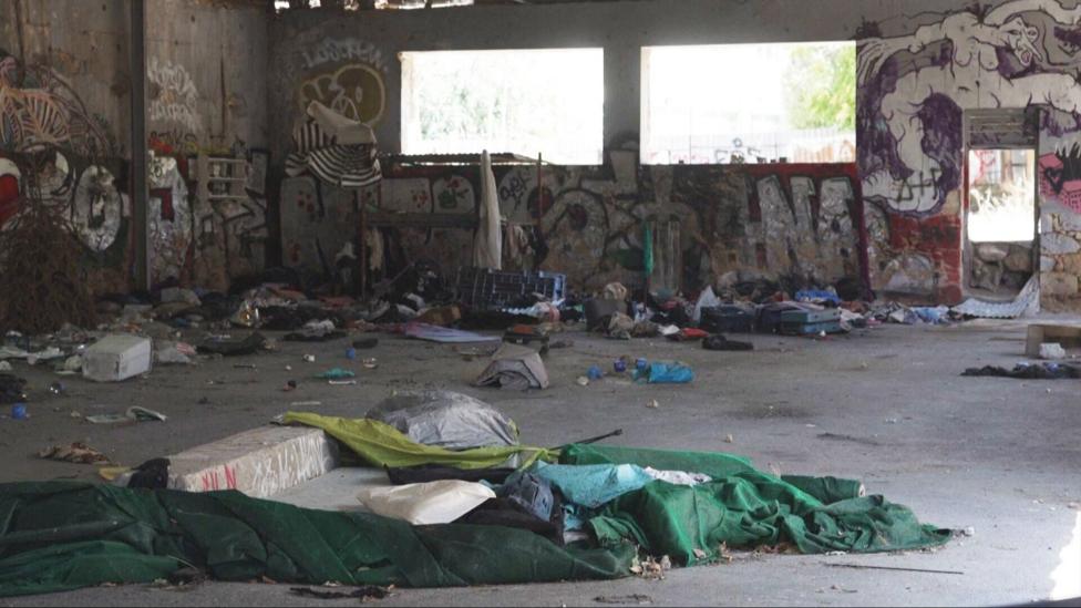 The inside of a neglected warehouse. The walls are graffitied and rubbish is strewn on the concrete floor. Bright light streams in through the window. 