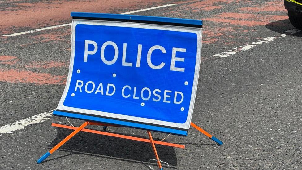 A blue sign with white writing. The writing says 'Police Road Closed'. The sign sits on orange legs on a road which has white and red markings