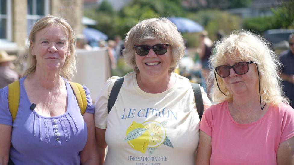 Lynne Sawyer, Debbie Saxby and Carol Alder, who are standing outside Westleton Village Hall. They are all smiling. Lynne is wearing a purple T-shirt, Debbie a white T-shirt and Carol a pink T-shirt.