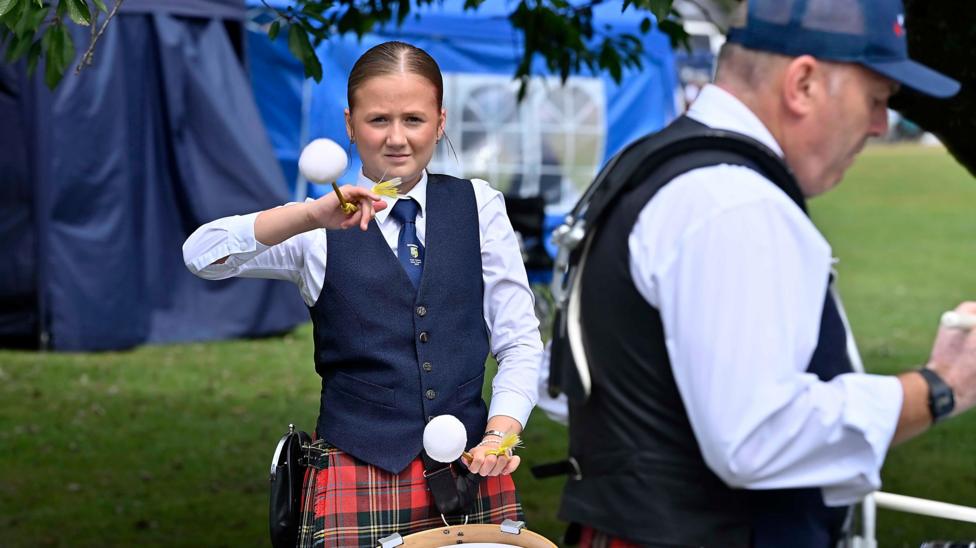 A young woman is playing drums. She's wearing a red checked kilt and a navy waistcoat. A ,am wearing a cap is next to her.