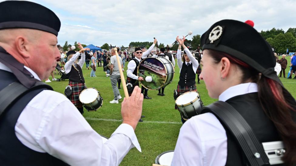 A man and woman are in the foreground. They're looking at each other and their backs are to the camera. They're holding drumsticks and drums. In front of them are drummers playing their instruments. Spectators are watching.