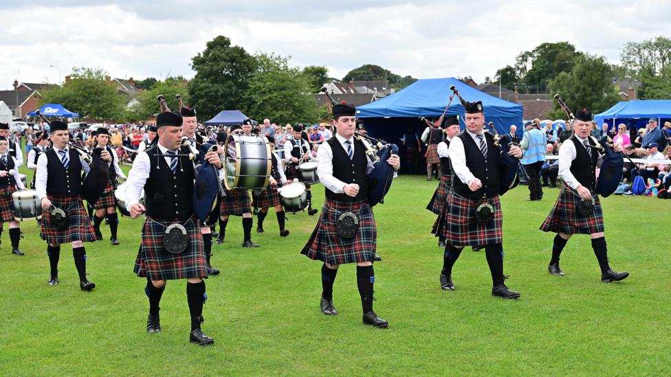 A group are holding pipes and drums and walking across grass. Spectators are watching. The sky is cloudy. Marquees are up.