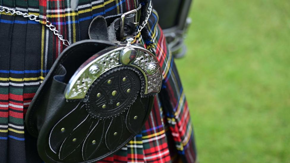 A close up of a black leather pouch with silver trimmings that is attached to a kilt. Grass is behind the person.