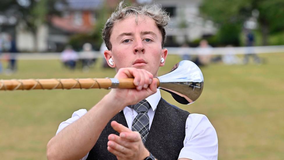 A young man is holding a baton. He has earpods in. He's wearing a black waistcoat with a checked tie and a white shirt.