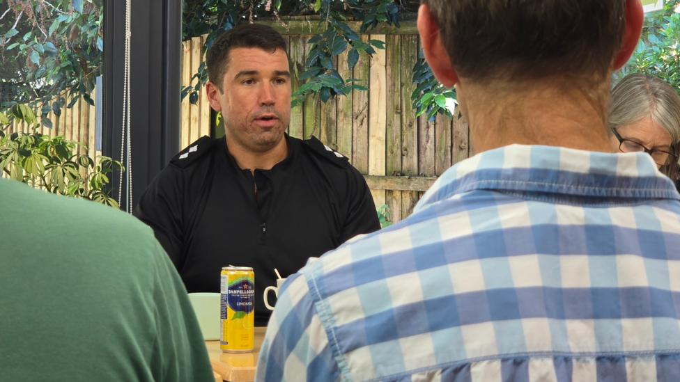 A police officer speaking to members of the public. He's wearing a black South Wales Police uniform. There is a man looking at him in a blue and white checkered shirt. There's a green shoulder by him. There's also a woman with grey hair and glasses sat next to the officer. 