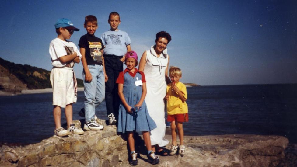 Anton central in a blue t shirt. He is flanked by three boys, a girl and a woman. They are stood on a rock with the sea and coast behind them.
