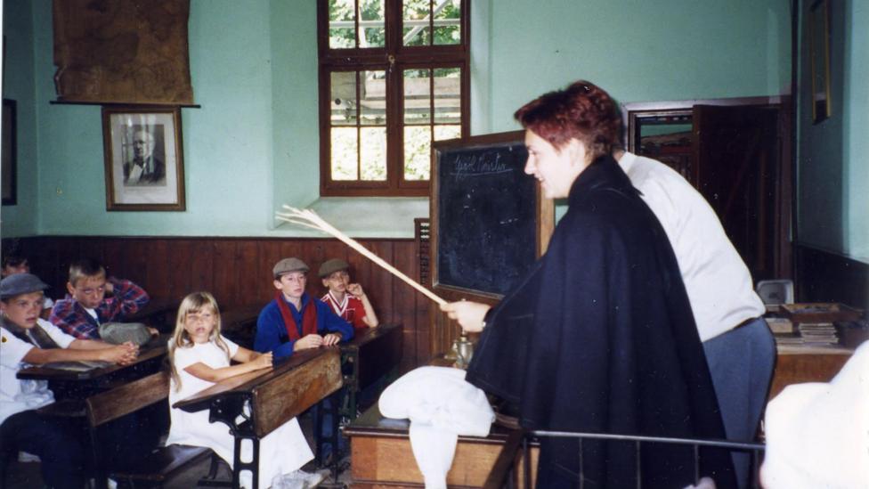 A woman stands at the front of an old classroom with a chalkboard behind her. Children, including Anton, sit at tables watching her.