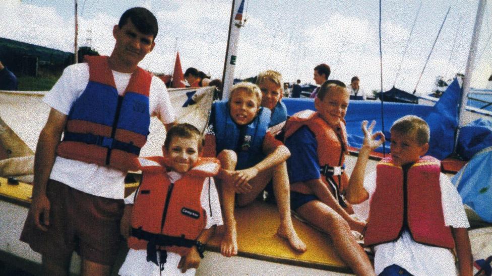 Anton sits on a boat with the Amoss family. They are all wearing life jackets and there are people stood behind them with the masts sticking in the air from other boats.