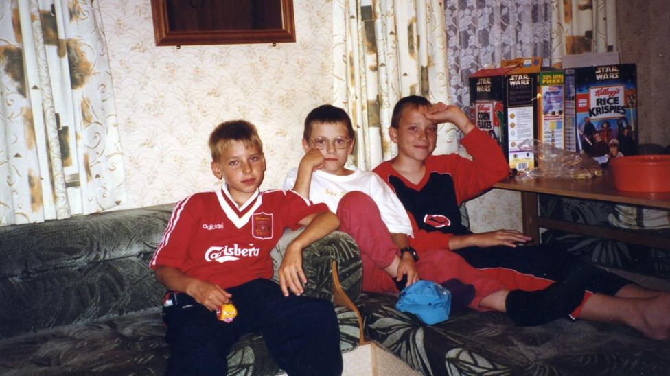 Dimtry on the left wearing an old Liverpool football top. Two boys in pyjamas sit on the right of him. There is a table on the right with cereal boxes.