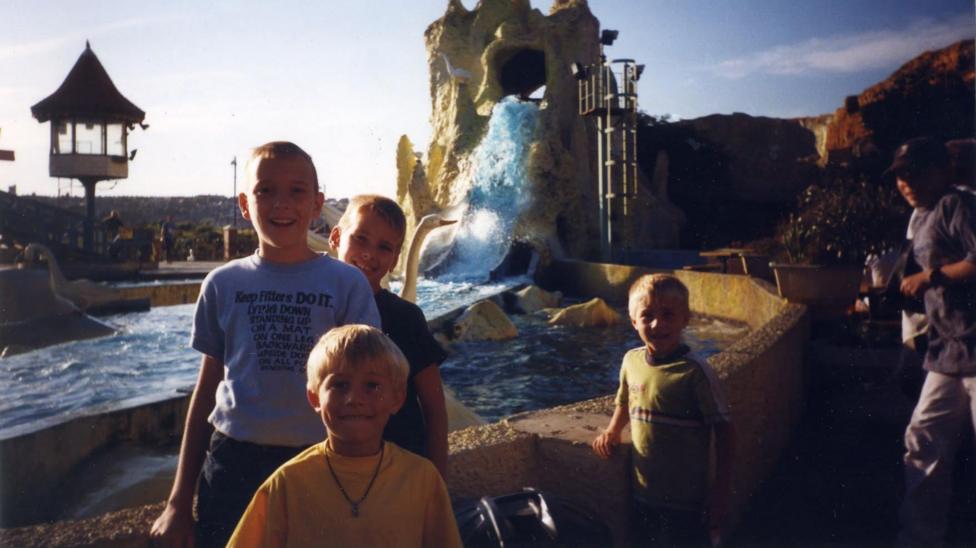 Anton on the left with a grey t shirt. He is stood next next to three boys and there is a attraction behind them at a waterpark. 