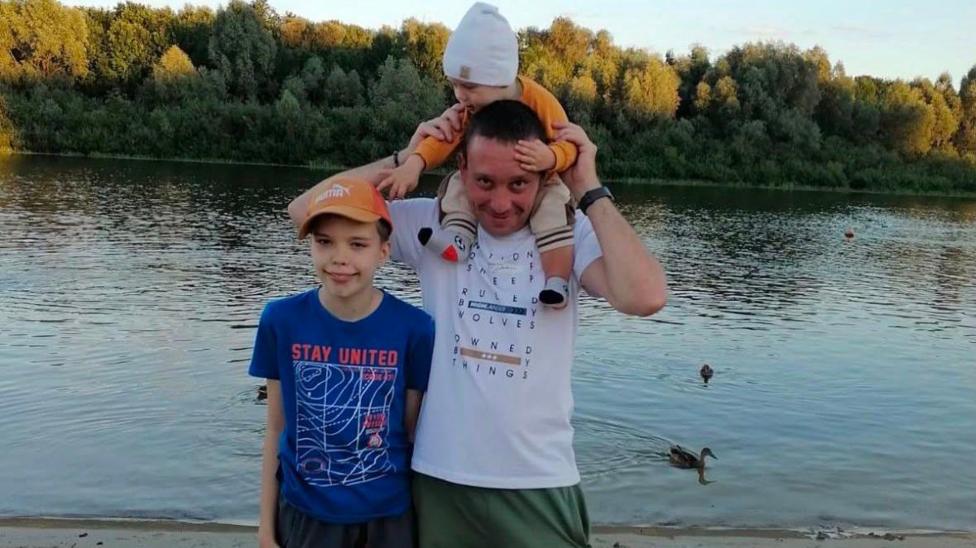 Anton in a white t shirt stood with two boys. One is on his shoulders holding onto his head. The other is standing by his side with a blue t shirt on.