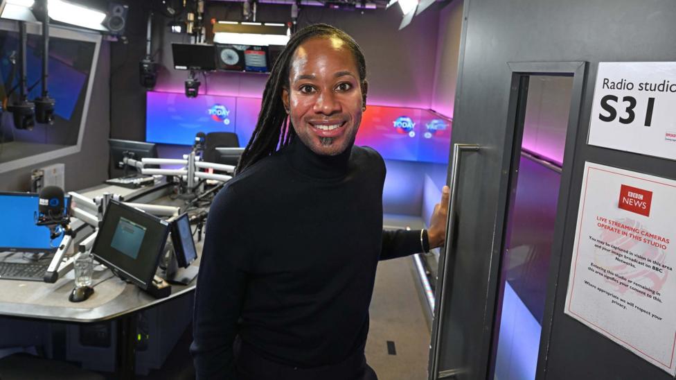Headshot showing Professor Jason Arday looking into the camera smiling, standing inside the BBC's Today programme studio. He is a black man with long black locs, ear piercings, wearing a black polo neck.