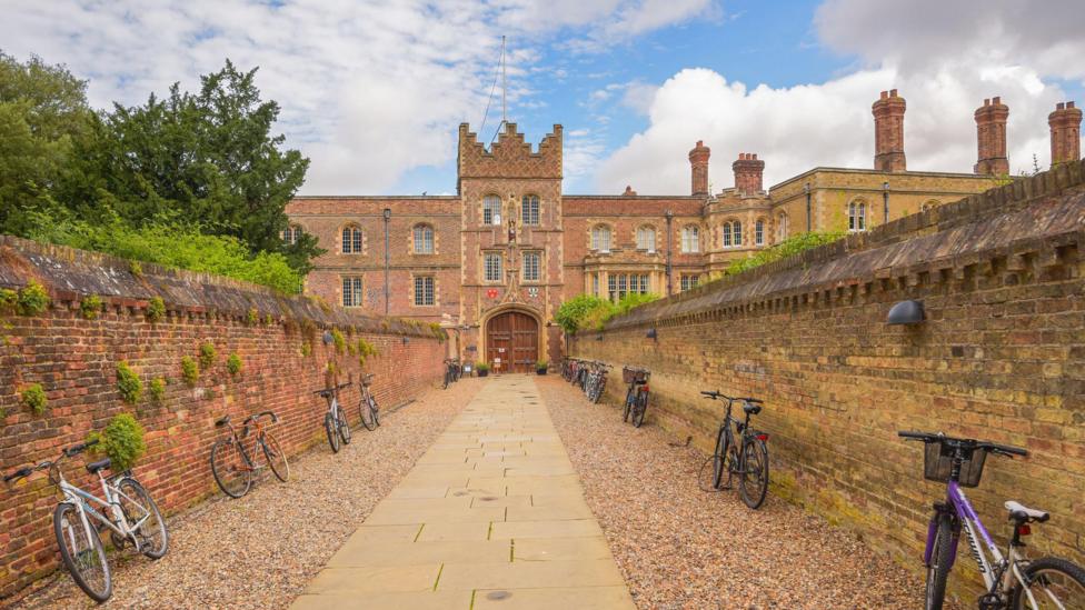 A wide shot of the pathway leading to Jesus College at Cambridge University on a sunny day. There are two brick walls on either side of a stone-paved path with pebbles on either side. There are eight bikes symmetrically-placed lined up neatly against the walls