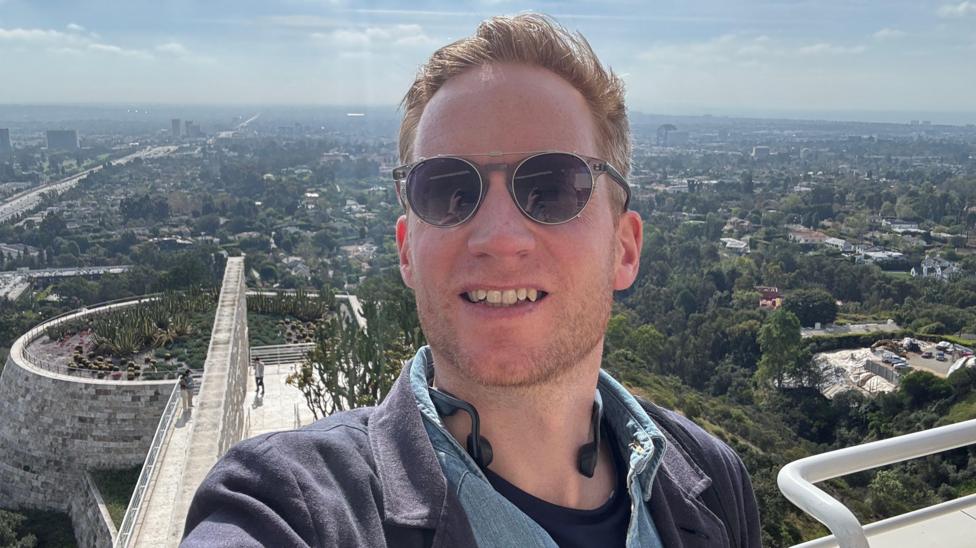 Kieran wearing sunglasses taking a selfie from an elevated outdoor terrace, with a sprawling cityscape and green hills in the background. A curved stone walkway and landscaped rooftop garden are visible below under a bright, hazy sky.